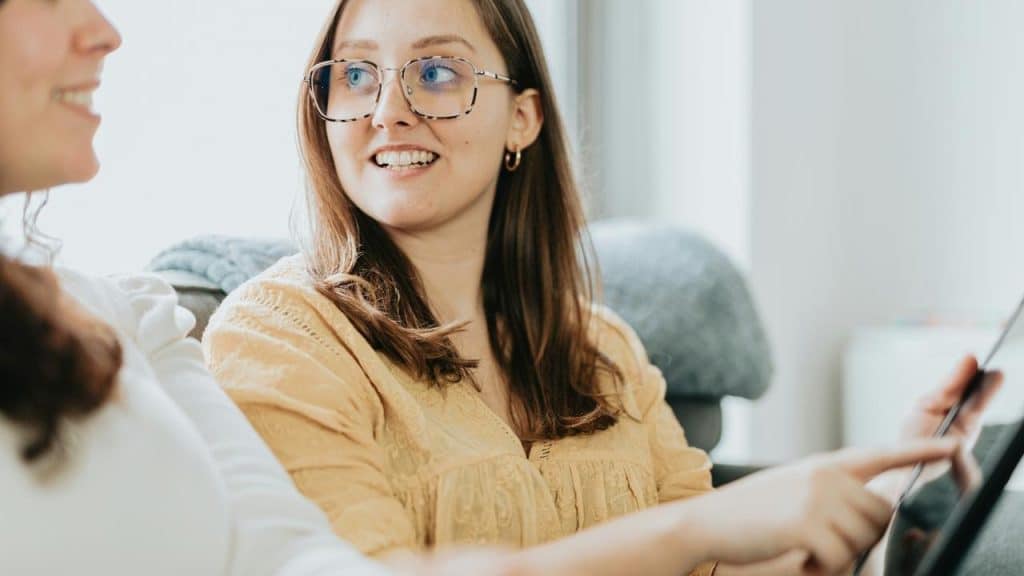 A woman wearing glasses talking with another person while holding a tablet indoors.