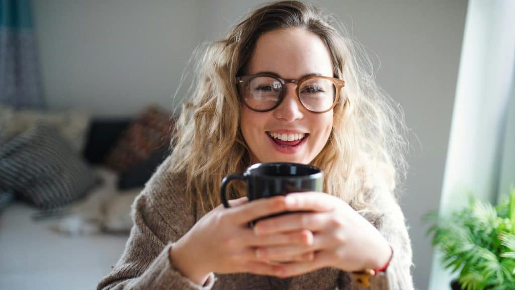 A smiling woman wearing glasses holding a mug indoors.