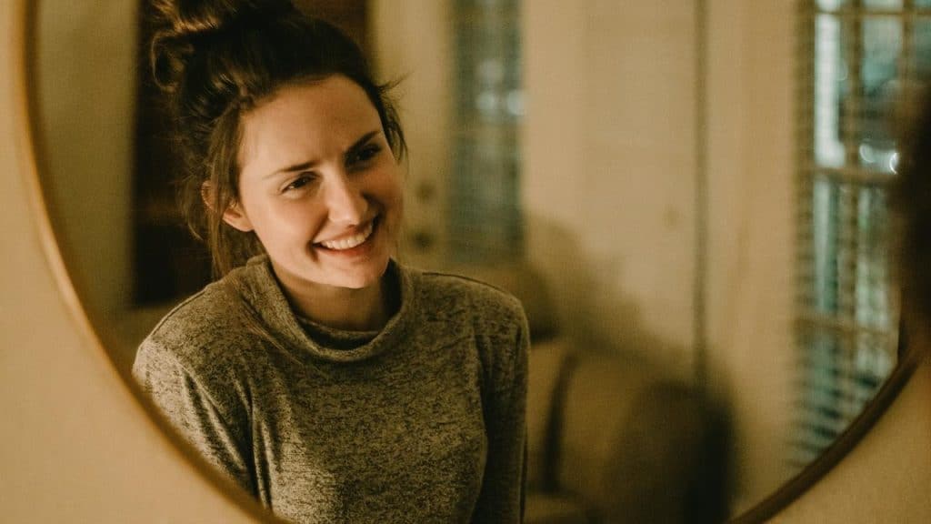 A smiling woman reflected in a mirror indoors.