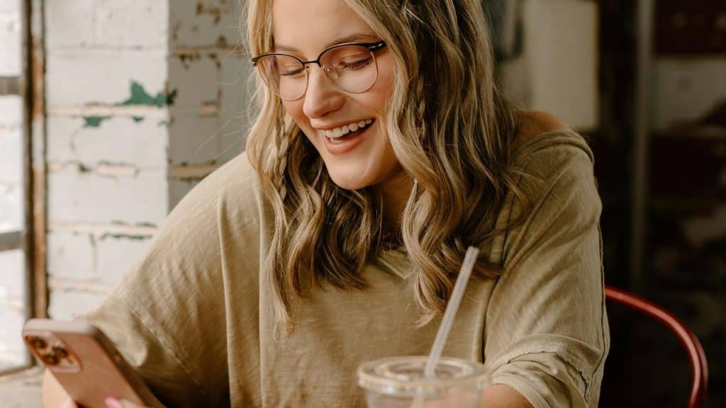 A woman wearing glasses smiling while looking at her phone indoors.