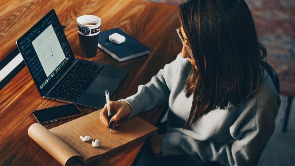 A woman working at a desk, writing notes beside a laptop and coffee.