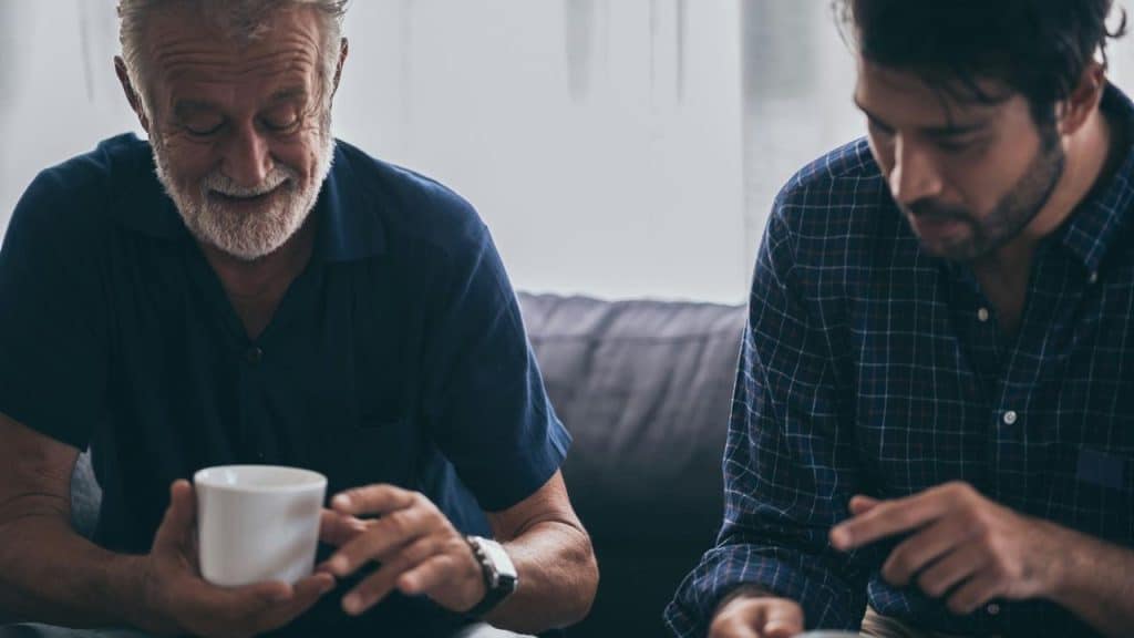 An older man holding a coffee while sitting with a younger man in conversation.