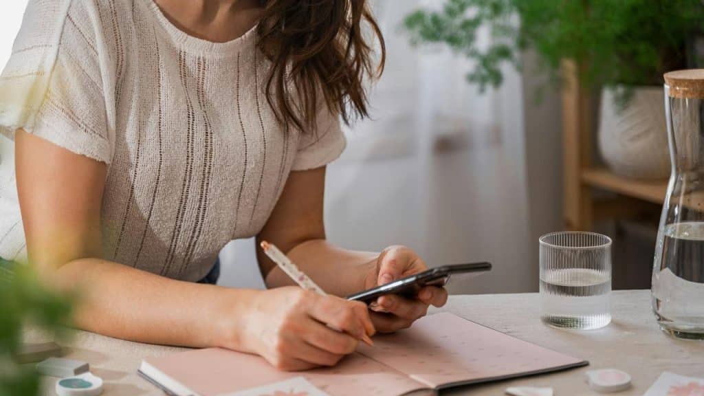 A person writing in a planner while checking a smartphone at a table.