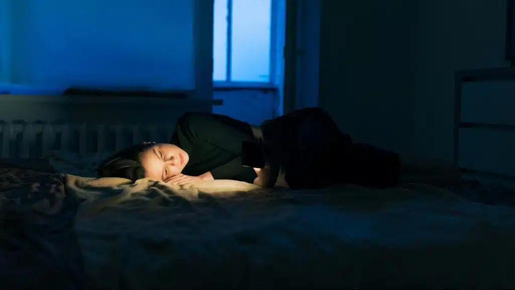 A woman lying on a bed at night, softly lit by the glow of her phone in a dark room.