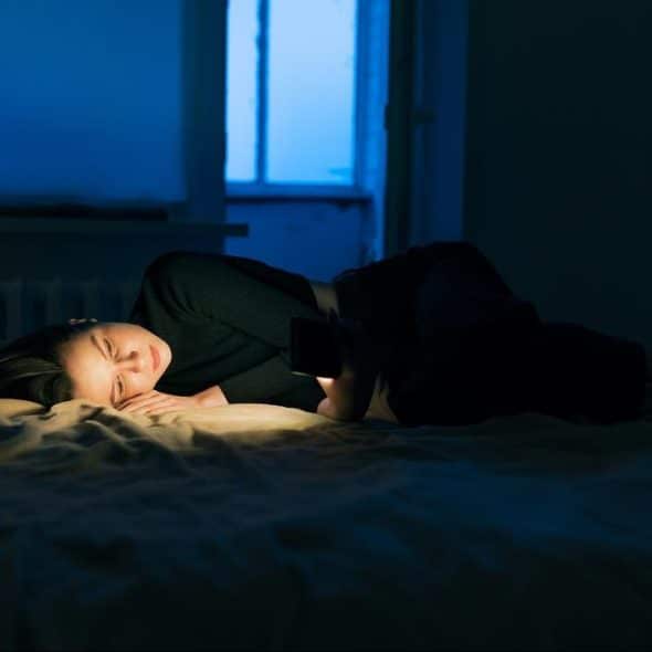 A woman lying on a bed at night, softly lit by the glow of her phone in a dark room.