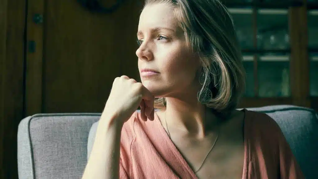 A woman sitting on a couch, gazing thoughtfully toward a window in soft daylight.