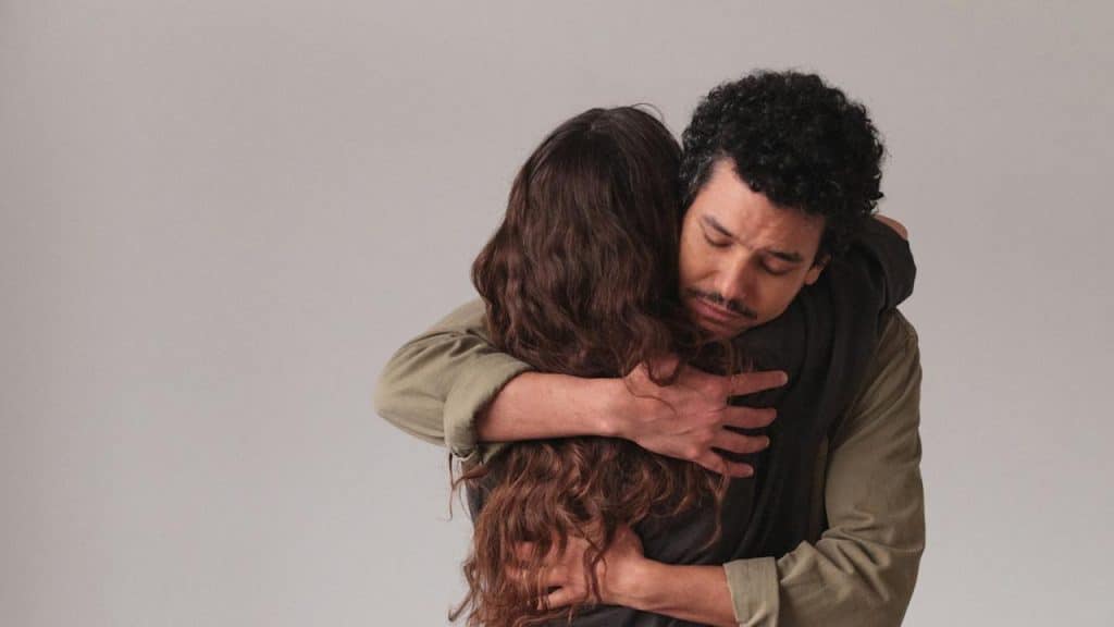 Man with a mustache hugging a woman from behind with a serious expression against a plain background.