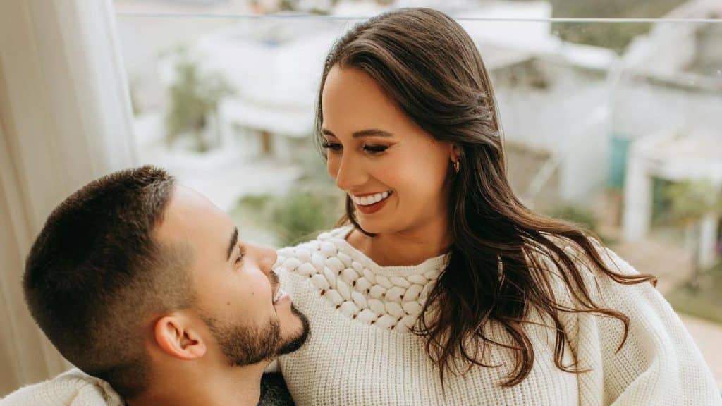 Close-up of a smiling couple looking at each other, with the woman wearing a white knit sweater.