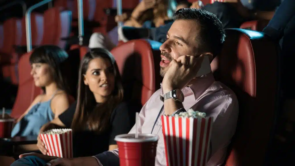 A man is talking on the phone while his jealous girlfriend is looking at him with anger while they are sitting in a cinema.