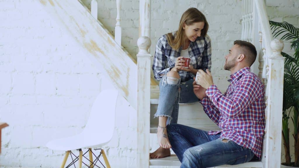 A man and a woman having coffee while sitting on stairs together at home.