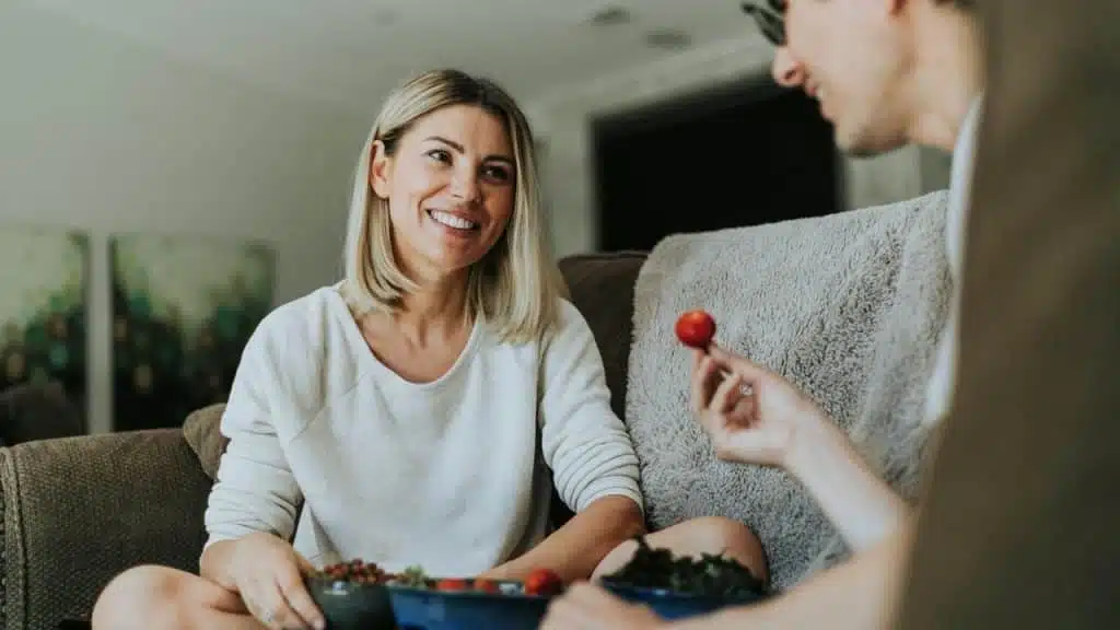 A woman smiling at a companion while sharing food on a couch indoors.