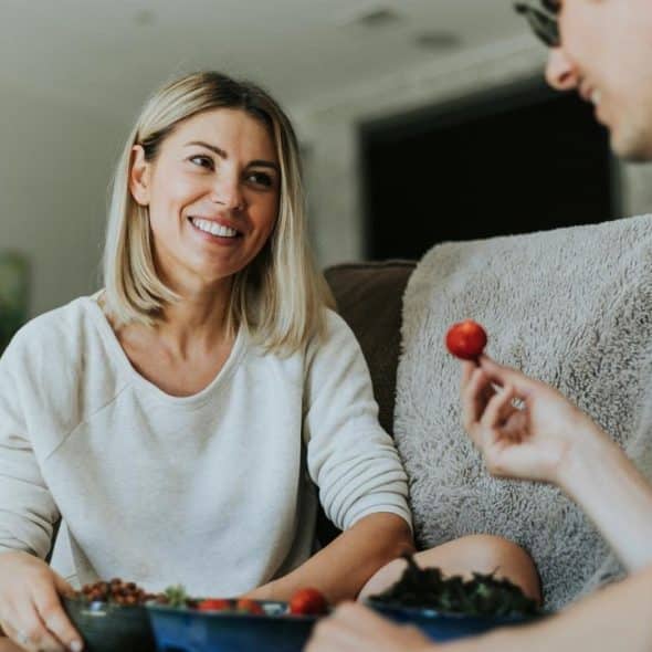 A woman smiling at a companion while sharing food on a couch indoors.