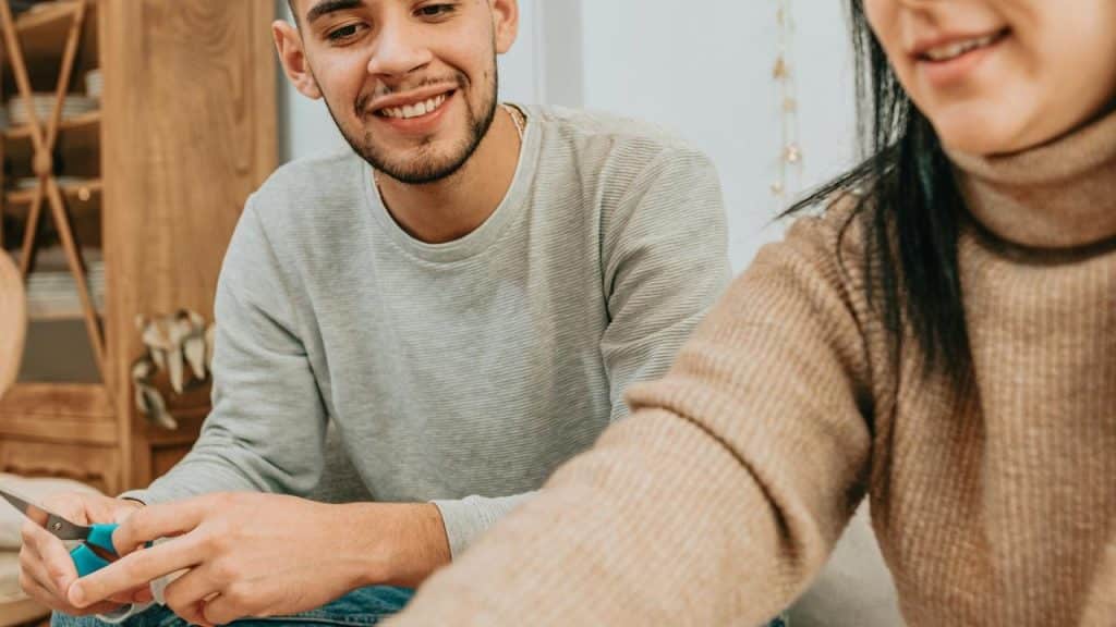 A smiling man sitting indoors while preparing food with another person.
