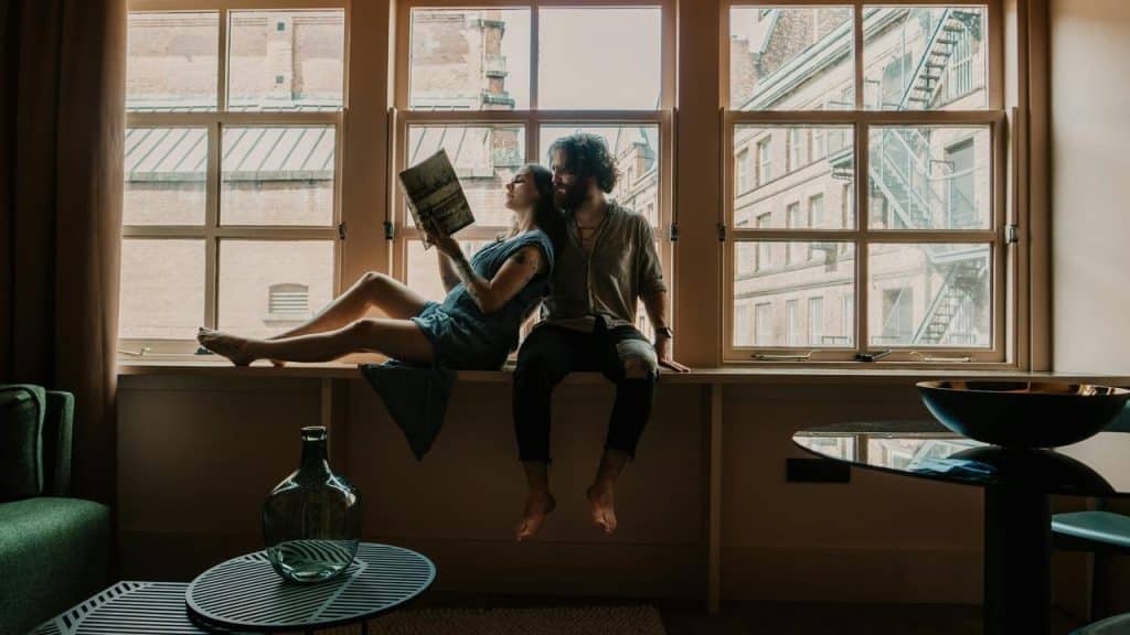 A couple sitting by a window indoors while one reads a book.