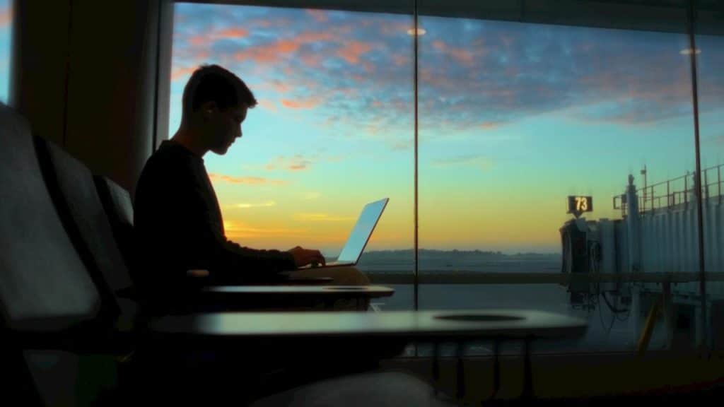A person working on a laptop at an airport gate during a colorful sunset.