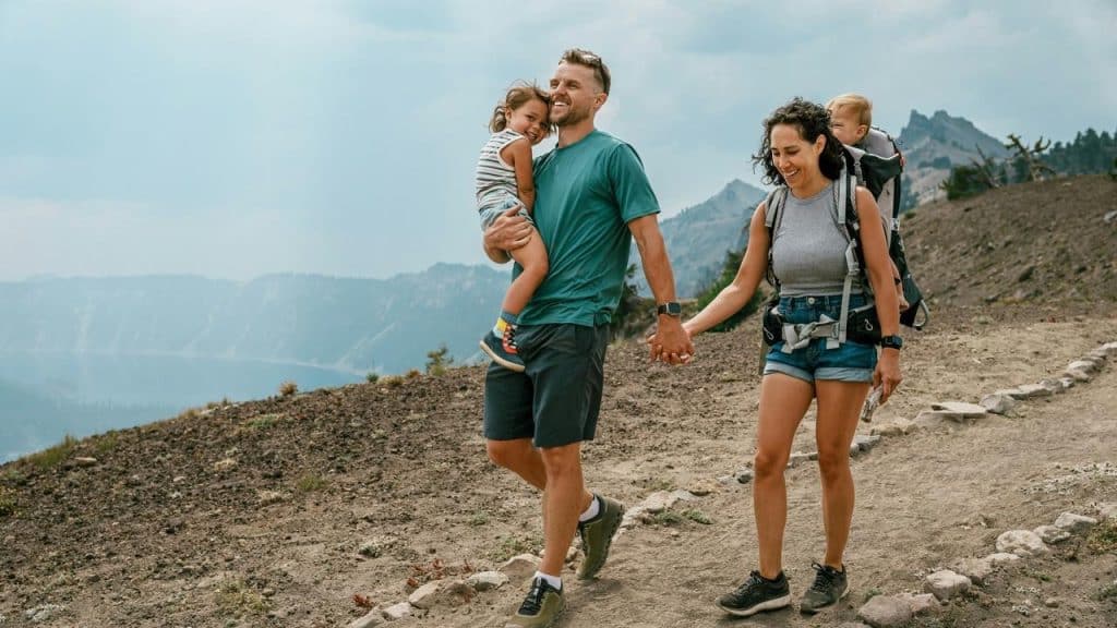 A family hiking on a mountain trail, with parents holding hands and carrying young children.