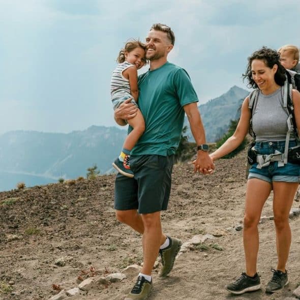 A family hiking on a mountain trail, with parents holding hands and carrying young children.