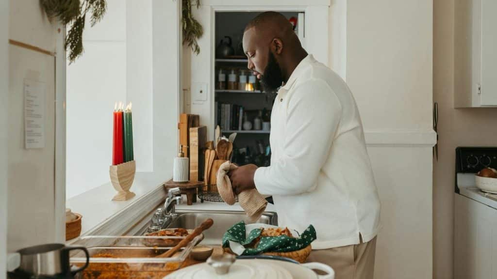 A man washing dishes at a kitchen sink beside lit candles and prepared food.