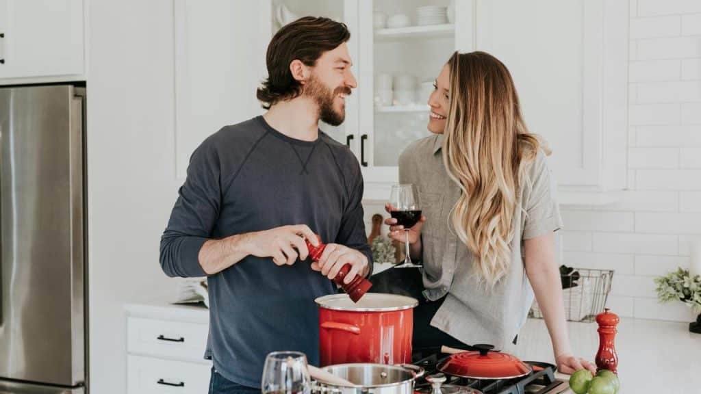 A couple smiling and cooking together in a bright kitchen while sharing a drink.