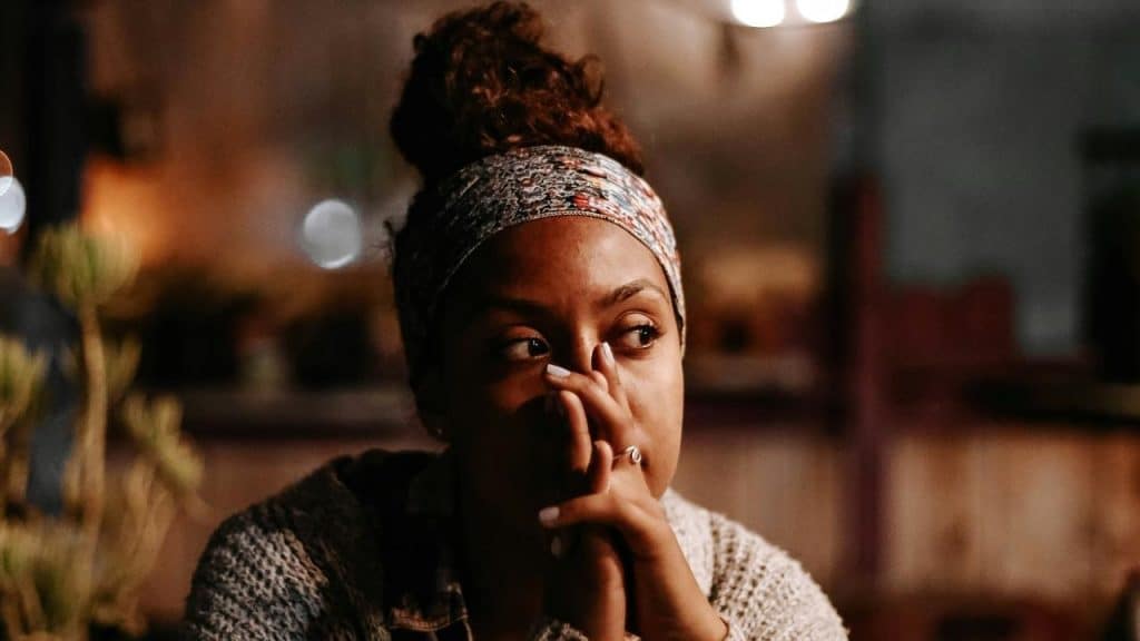 A thoughtful woman sitting indoors at night with her hands near her face.