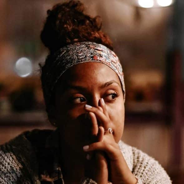 A thoughtful woman sitting indoors at night with her hands near her face.