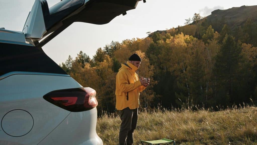 A man in a yellow jacket standing by an open car trunk in a grassy autumn landscape.