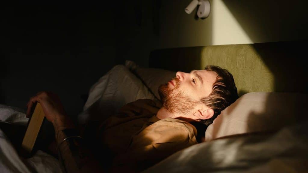 A man lying in bed, holding a book as warm light falls across his face.