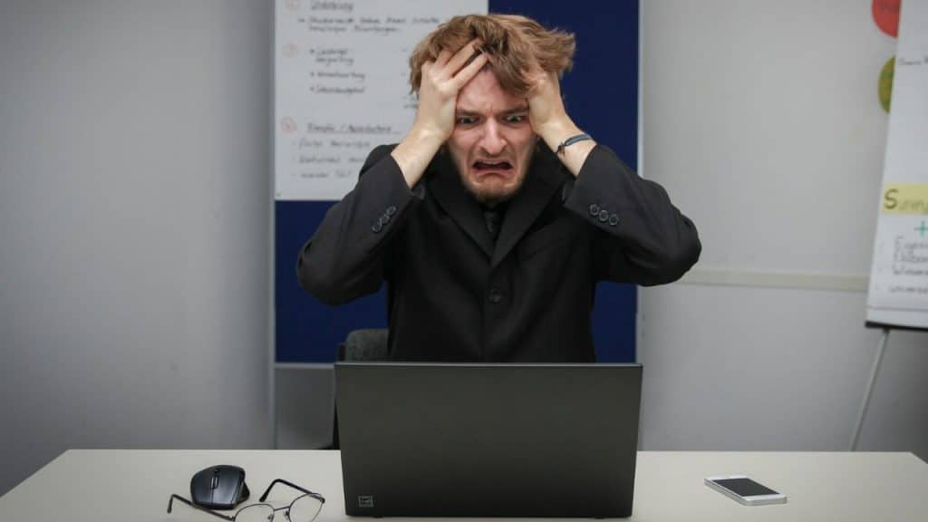 A man in a black suit sits at a desk, clutching his head behind a laptop.