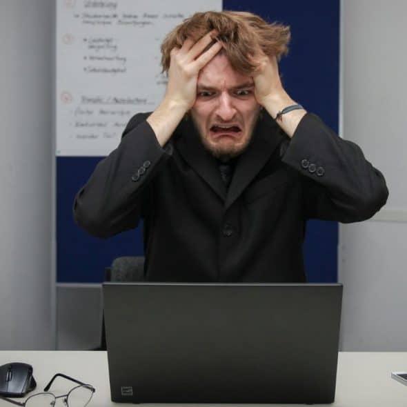 A man in a black suit sits at a desk, clutching his head behind a laptop.
