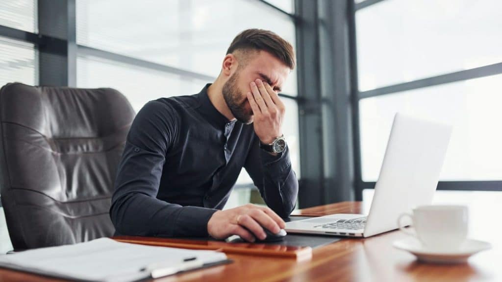 A man in a dark shirt sits at a desk, rubbing his eyes while using a laptop.