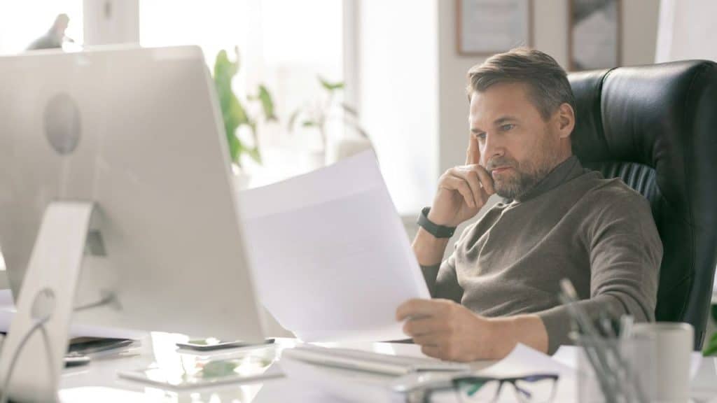 A man in a brown sweater sits at a desk, looking at a paper with a neutral expression.
