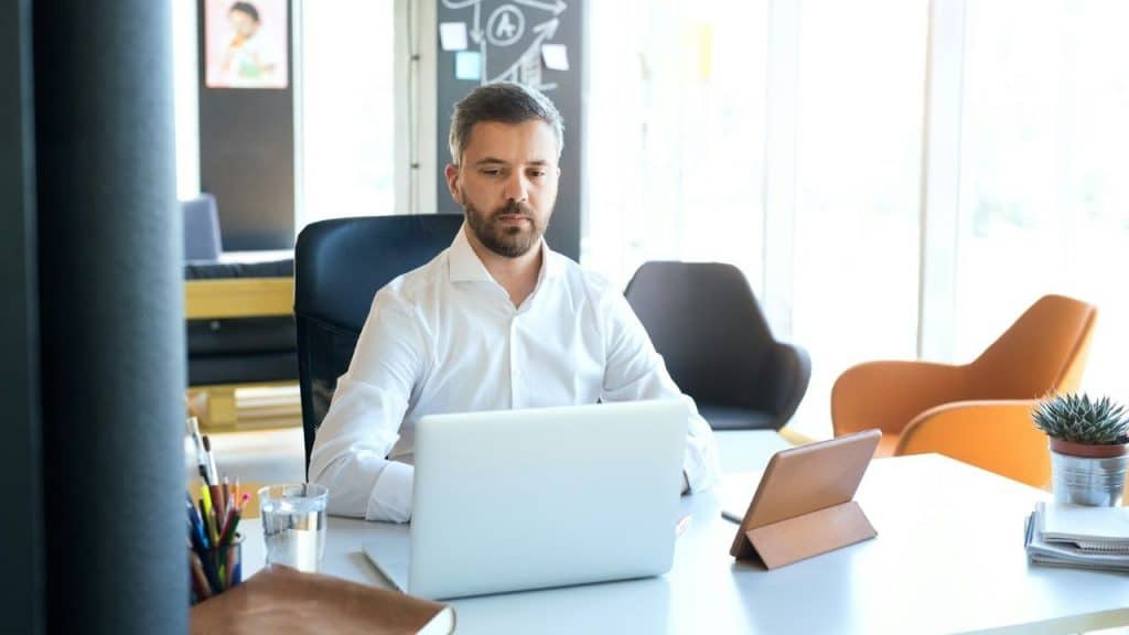 A man in a white shirt sits at a bright desk, looking at a laptop screen.