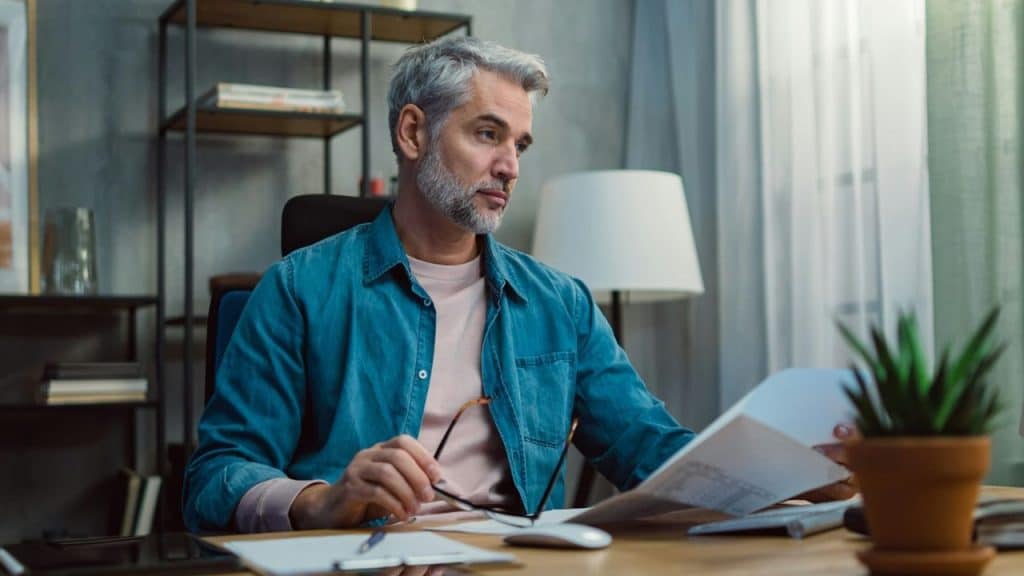 A middle-aged man with grey hair sits at a desk, looking thoughtfully at a document.