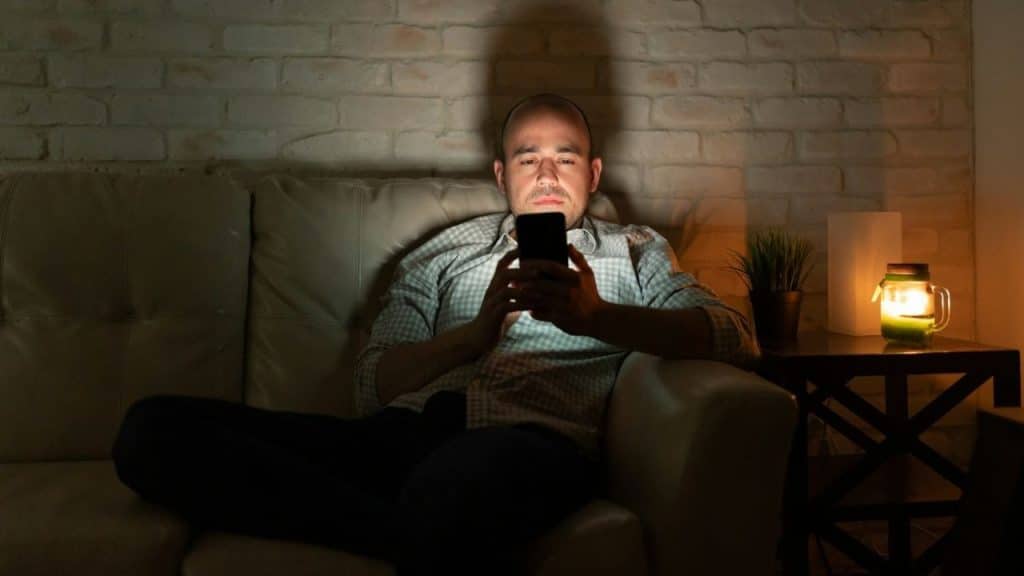 A man sits on a dark sofa at night, illuminated by his glowing smartphone.