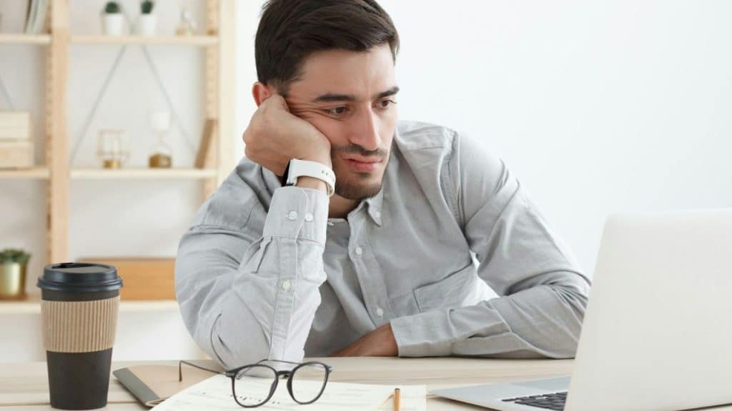 A man in a light grey shirt sits at a desk, leaning his chin on one hand.