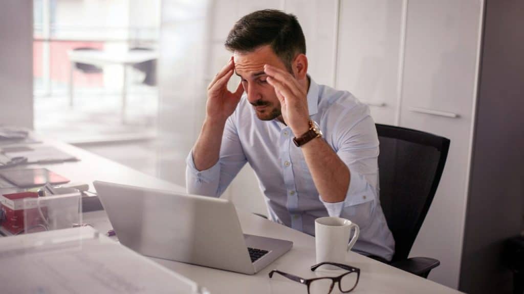 A man in a light blue shirt sits at a desk, touching his temples behind a laptop.