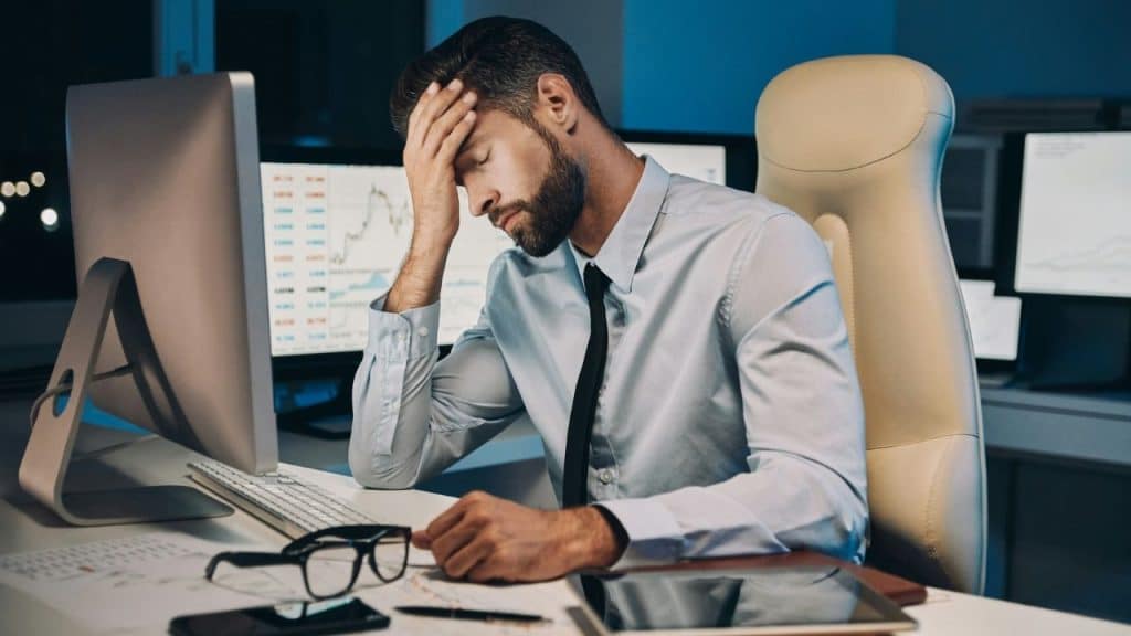 A man in a light blue shirt sits at a desk, leaning his forehead on one hand.