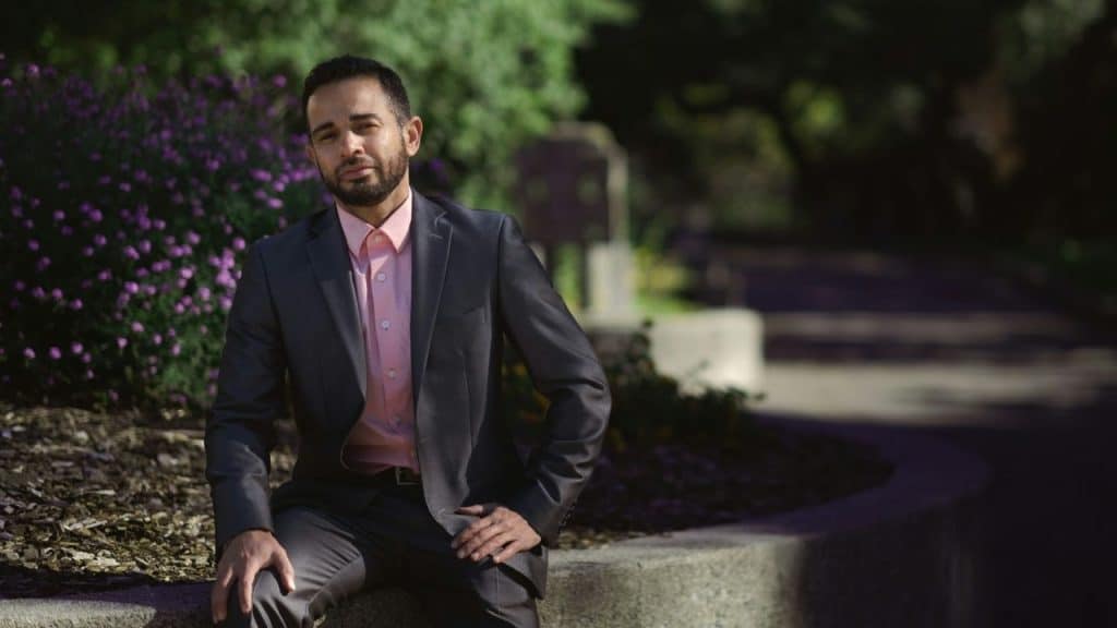 A man in a suit sits on a stone ledge in a park with a neutral expression.