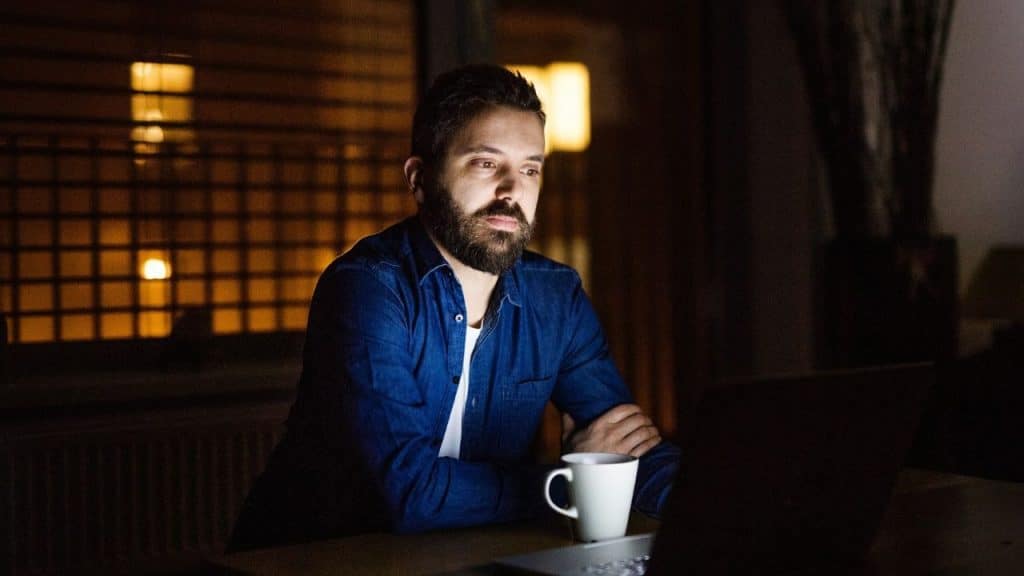 A man with a beard sits at a dark desk with a white mug and laptop.