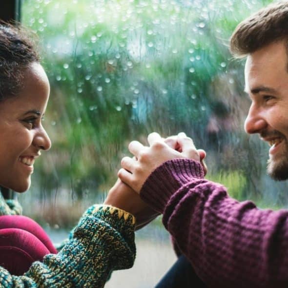 A man and woman sit by a rainy window, smiling and holding hands while facing each other.