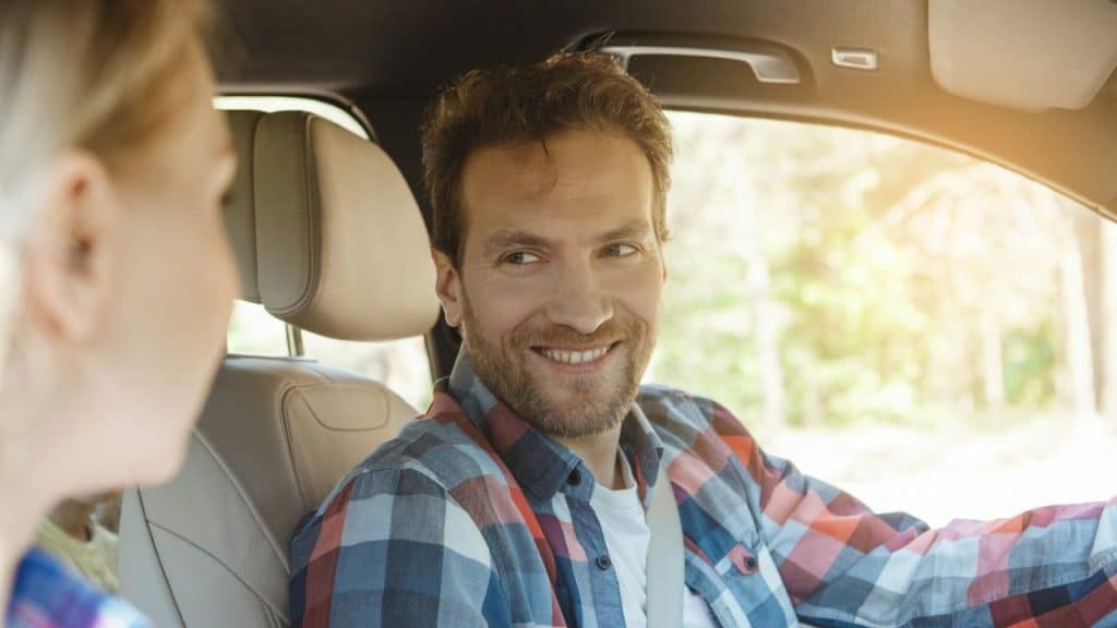 A man in a plaid shirt smiles while looking at a passenger inside a car.