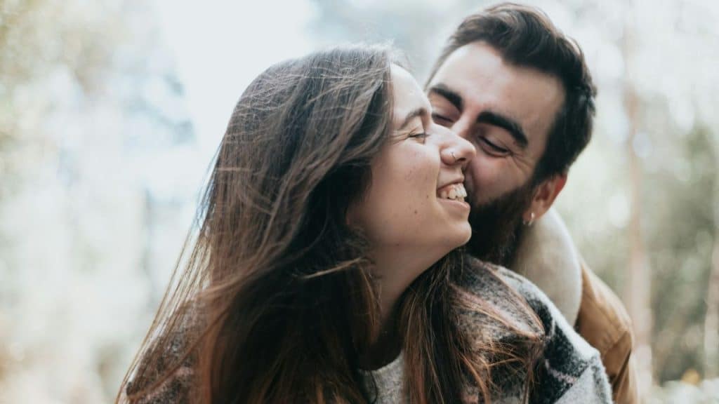 A man with a beard leans in to kiss a smiling woman's cheek outdoors.