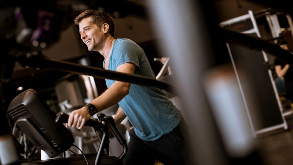 A man in a blue shirt smiles while exercising on an elliptical machine in a gym.
