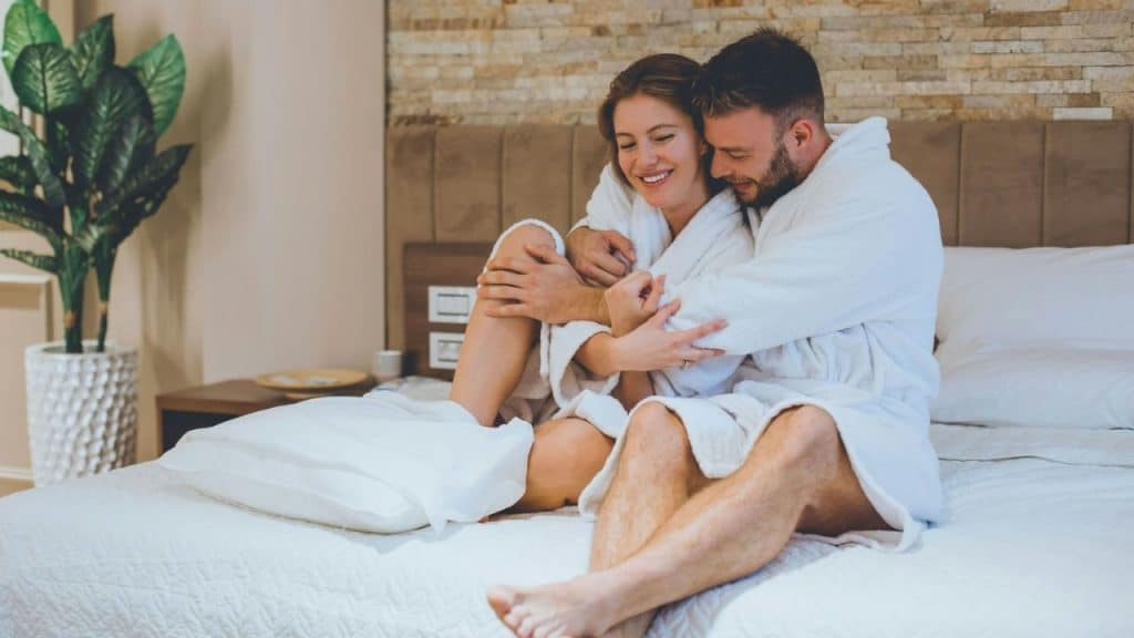 A man and woman in white bathrobes sit on a bed together while smiling and hugging.