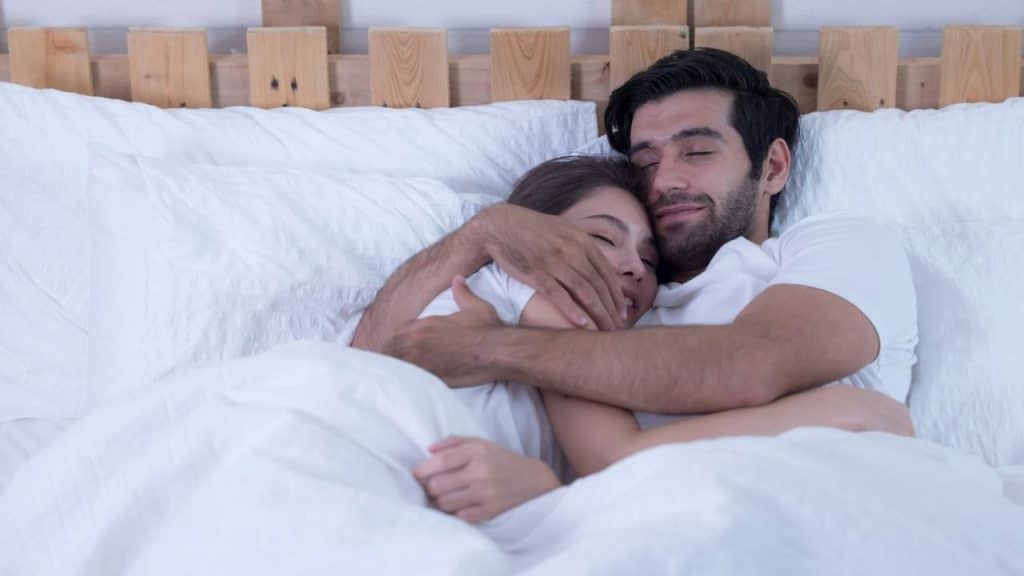 A man and woman lie in bed together, smiling with their eyes closed while hugging.