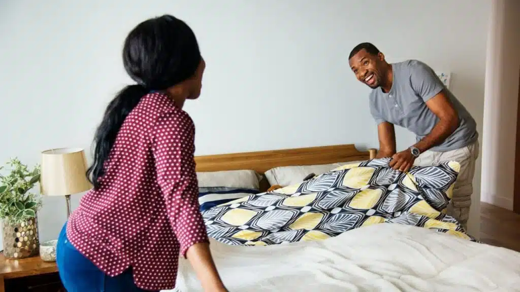 A man and woman smile at each other while pulling a patterned duvet over a bed.