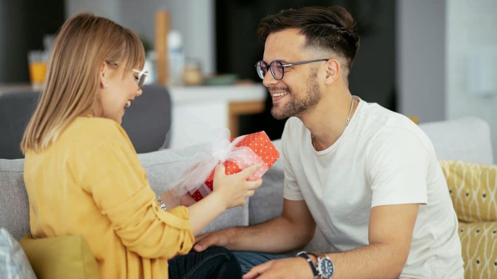 A man in glasses smiles while giving a wrapped gift to a woman on a couch.