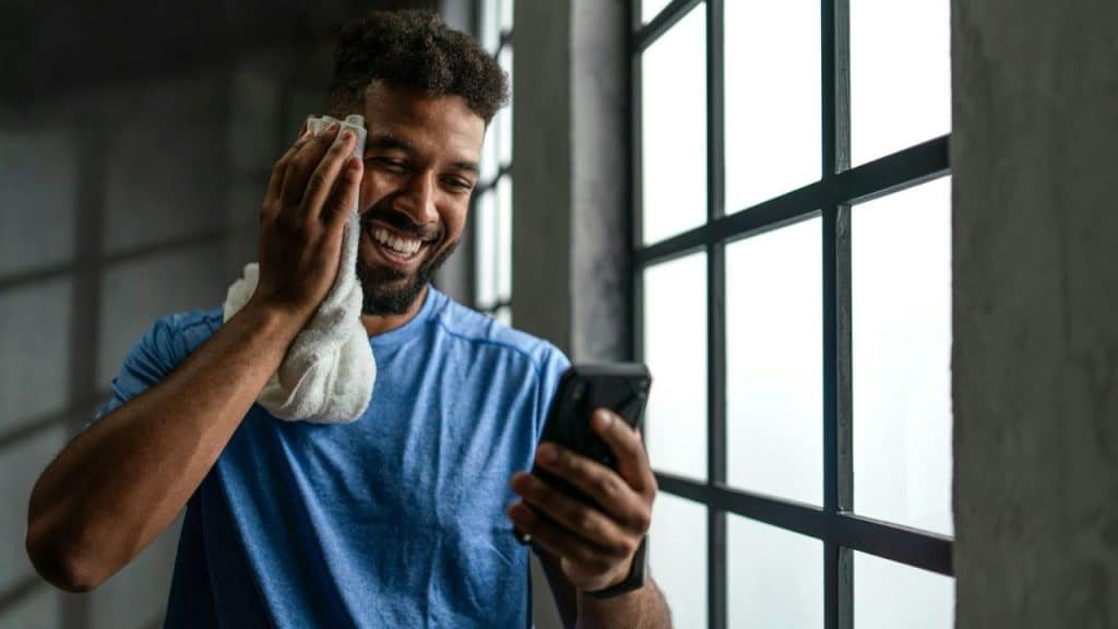 A man in a blue shirt smiles while holding a phone and wiping his face.