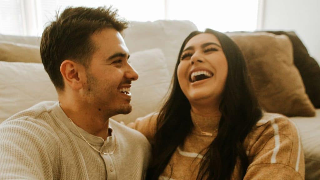 A man and woman sit on a couch together, smiling and laughing with each other.