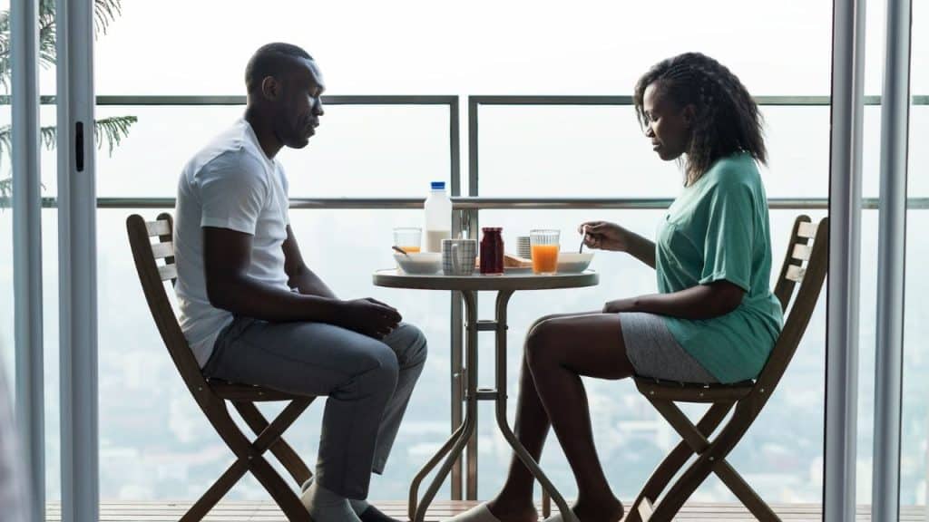A man and woman sit at a small balcony table eating breakfast in silence.